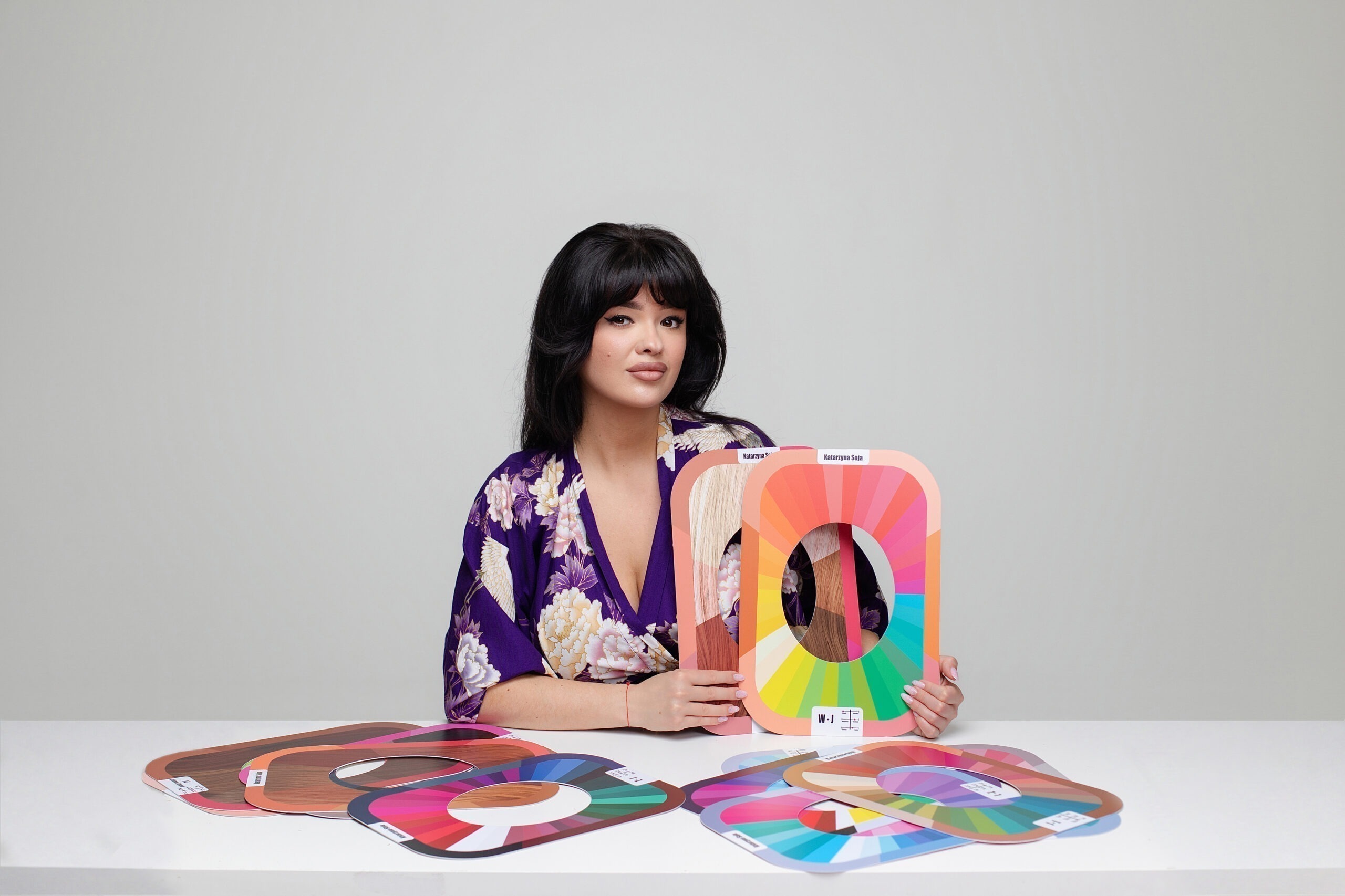 Woman in a purple floral kimono holds a large color wheel swatch card, with many color swatches laid out on the table in front of her, in a studio setting.